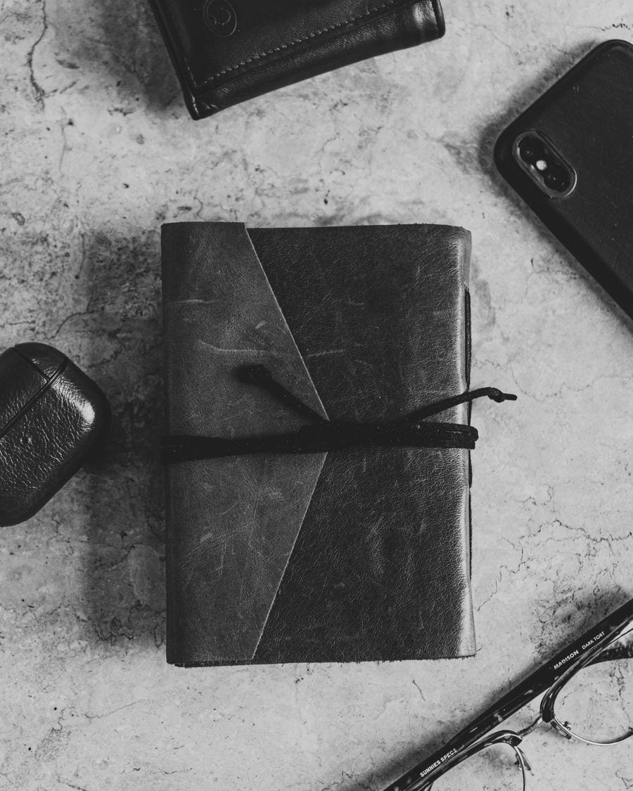 A leather notebook, pen, and loose papers laid out on a desk, in black and white
